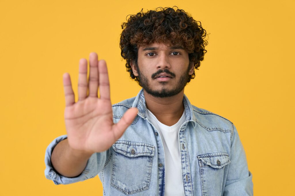 Young indian man showing stop hand gesture isolated on yellow background.