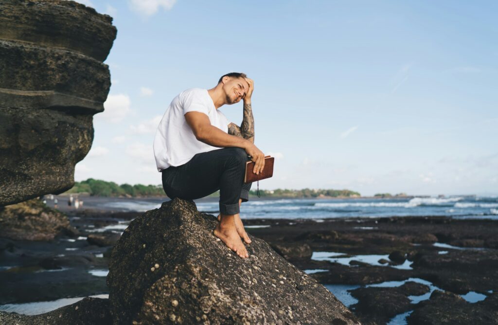 Young man sitting on rock at shore and thinking over interesting book